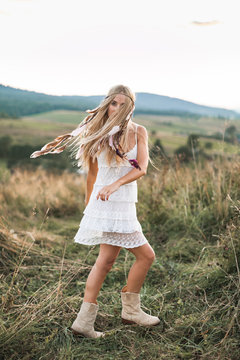 Beautiful young boho hippie girl in white dress at sunset, smiling and having fun. Feathers in hair, bohemia boho style
