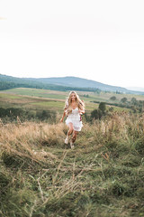 Naklejka na ściany i meble happy smiling woman with long blonde hair wearing white boho hippie dress having fun while running in field. Feathers in hair, boho style
