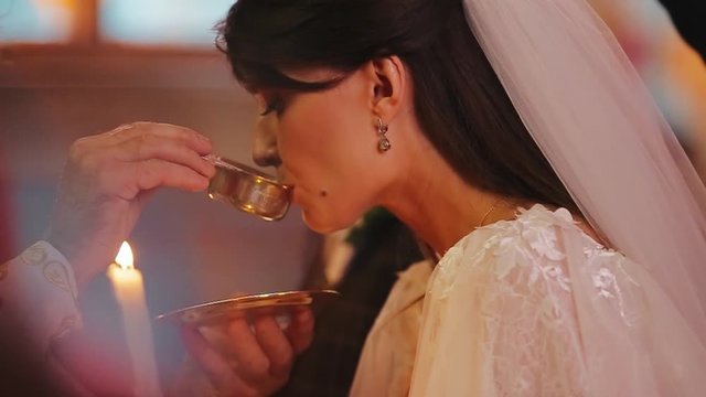 Beautiful bride in luxurious white dress drinks sacred water in gilded cup in the hands of priest. Christian tradition of church wedding in Ukraine. European custom for believers in love at ceremony.