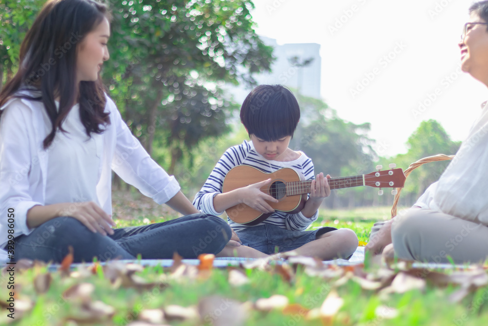 Poster Happy Asian family has leisure in the park on the weekend or holiday. Mother teach son to play guitar. Grandmother enjoy this beautiful moment of love. Family generation relationship or bonding photo