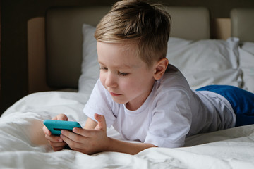 Naklejka premium Cute little boy toddler playing mobile game, lying on a bed and holding smartphone