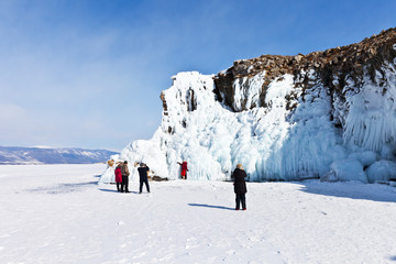 Winter Lake Baikal. Happy tourists take pictures on the background of the beautiful icy cliffs of Olkhon Island on a sunny day. Traveling on a frozen lake