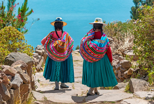 Two indigenous Quechua women in traditional clothes walking down the path to the harbor of Isla Taquile (Taquile Island) with the Titicaca Lake in the background, Peru.