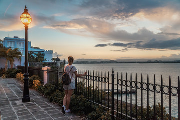 Woman is standing next to the gates at the look out point in old San Juan, Puerto Rico. She is watching sunset. Beautiful and colorful lights are on the background. Sea is calm and walled city also. - obrazy, fototapety, plakaty
