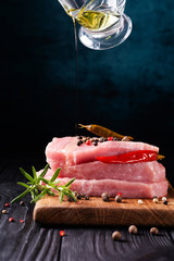 Stack of steaks of fresh raw pork meat decorated with chili peppers and rosemary with chef's action pouring olive oil on a dark background. Copy space.