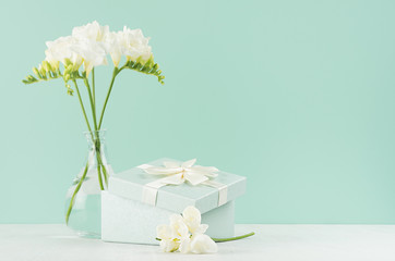 Fresh delicate white flowers freesia and festive opened square gift box with ribbon and knot in green mint menthe interior on white wood board.