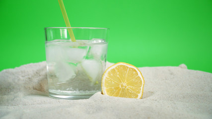 glass with ice and soda water and lemon on the sand on a green background