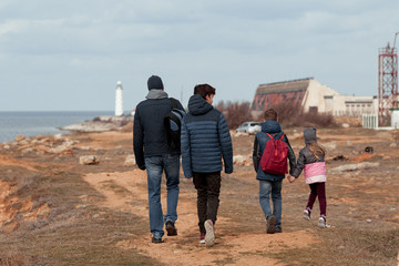 A father with two older sons and a small daughter walks along the seashore, the family goes to the lighthouse, spring, they are dressed in jackets