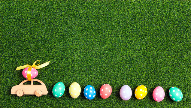 Wooden toy car with Easter egg on the roof and row of eggs on a green grass background.