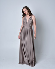 Fototapeta premium Portrait of a pretty brunette girl wearing a long silver evening gown, full length standing pose against a studio background.