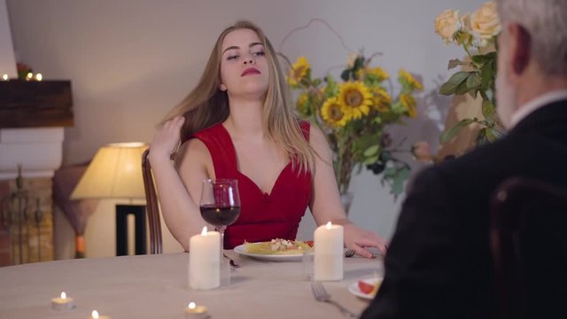 Portrait of confident young Caucasian woman in red seductive dress making money gesture. Grey-haired man in suit taking out wallet to pay prostitute. Lifestyle, escort service.
