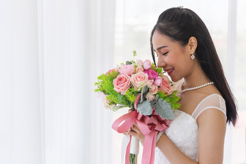 An Asian bride in a wedding dress is sitting smiling brightly on the bed in her hand holding a beautiful bouquet of flowers.