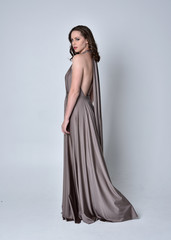  Portrait of a pretty brunette girl wearing a long silver evening gown, full length standing pose against a studio background.