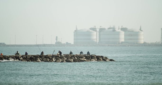 A group of fishermen at a breakwater fishing with gas storage tanks in the background