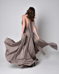  Portrait of a pretty brunette girl wearing a long silver evening gown, full length standing pose against a studio background.