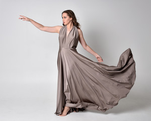  Portrait of a pretty brunette girl wearing a long silver evening gown, full length standing pose against a studio background.