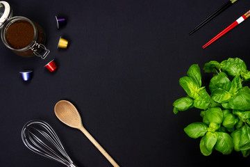 Top view of a whisk, a wooden spoon, a plant of green Basil, a pair of chopsticks, grinded coffee and four capsules of coffee . Dark and dramatic light. Black background. Studio set. Center copy space