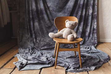 Loneliness concept. Beige teddy bear is sitting alone on a wooden chair in empty room. Vintage toys...