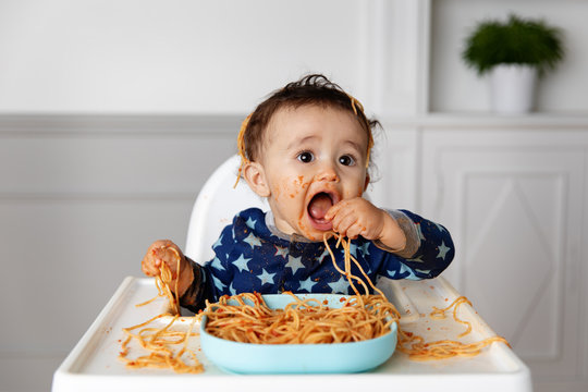 Funny baby in high chair eating spaghetti with his hands