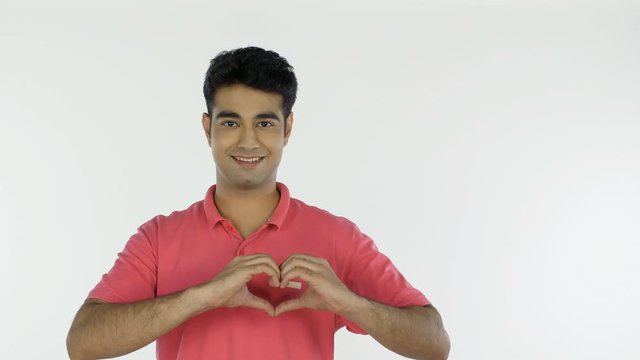 Attractive Indian man doing a hand heart gesture against the white background. Portrait of a happy young guy making a heart shape with his hands while looking into the camera - emotions concept