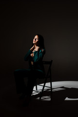 Studio photo of pretty brunette woman in twilight sitting on black chair. A ray of light hits her face.