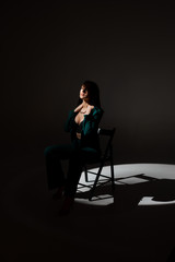 Studio photo of pretty brunette woman in twilight sitting on black chair. A ray of light hits her face.