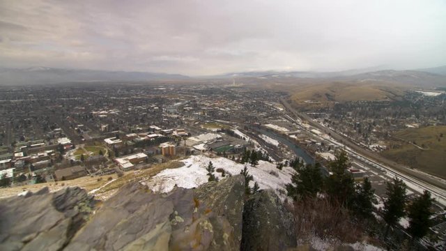 Slider shot right to left looking out over The University of Montana and Missoula.  Rock outcropping in foreground.  Snow down in valley.  Overcast sky.  Golden Light.