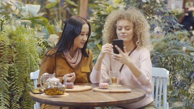 Two young attractive female friends taking selfie with smartphone, then looking at photo and discussing it while having tea and desserts in trendy cafe filled with green plants