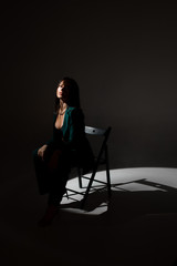 Obraz na płótnie Canvas Studio photo of pretty brunette woman in twilight sitting on black chair. A ray of light hits her face.