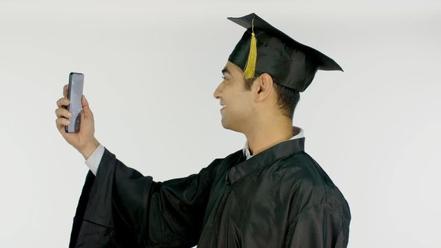 Indian student taking selfies with a smartphone during the convocation ceremony. Happy young graduate clicking pictures in graduation gown and cap against the white background - college lifestyle c...