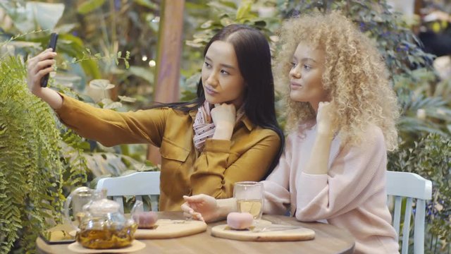 Young Asian woman using smartphone and taking selfie with her blonde female friend, then watching photo and discussing it while sitting at table in plant-filled cafe