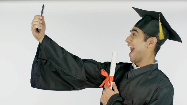 Male college grad happily taking selfies in a black robe and square academical cap. Young graduate excitedly clicking pictures with his smartphone while holding his graduation degree - white backgr...