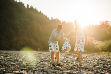 Mom, dad, daughter walking on stone near lake. The concept of summer holiday. Mother's, father's, baby's day. Family spending time together on nature. Family look. Sun light.