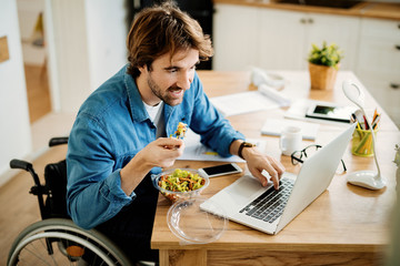 Smiling freelance worker in wheelchair working on laptop while eating at home.