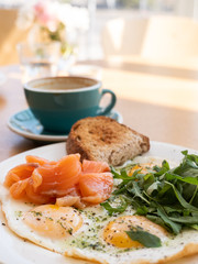 Tasty healthy breakfast: fried eggs, salmon, arugula and croutons on a white plate and  a mug of cappuccino on a wooden table.
