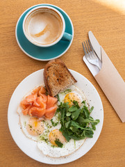 Tasty healthy breakfast: fried eggs, salmon, arugula and croutons on a white plate and  a mug of cappuccino on a wooden table.