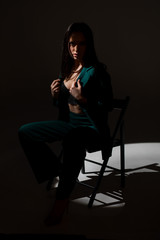 Studio photo of pretty brunette woman in twilight sitting on black chair. A ray of light hits her face.