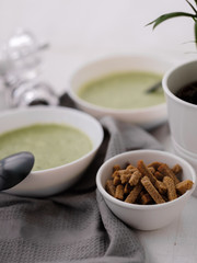 Spring detox broccoli green cream soup in white plates, bowl with crackers and stylish black cutlery on grey background. 
