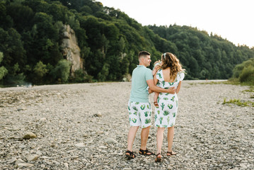 Mom, dad hugging daughter walking on stone near lake. The concept of summer holiday. Mother's, father's, baby's day. Family spending time together on nature. Family look. Sun light. Back view.