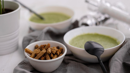 Spring detox broccoli green cream soup in white plates, bowl with crackers and stylish black cutlery on grey background. 