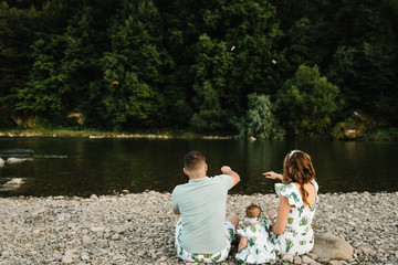 Family throwing stones into water. Mom, dad hugging daughter sitting on beach near lake. The concept of summer holiday. Mother's, father's, baby's day. Spending time together. Family look. Back view.