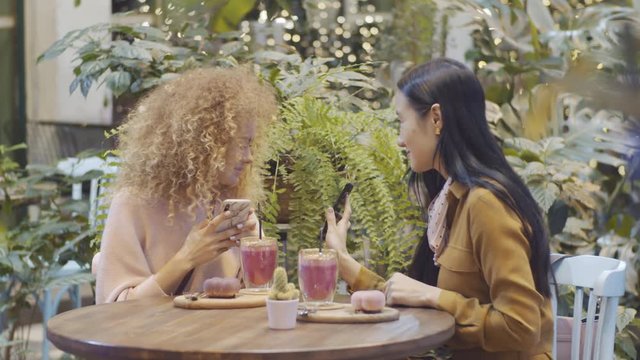 Two young Asian and Caucasian female friends photographing their smoothies and desserts with smartphones and discussing pictures while sitting at table near green plants in modern cafe
