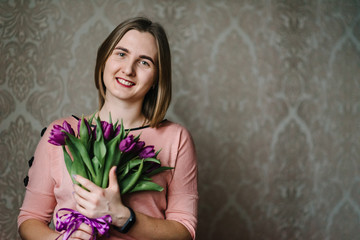 Happy women's day! Cute woman hold a bouquet of flowers at home. Beautiful girl in the dress with violet tulips in hands.