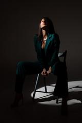 Studio photo of pretty brunette woman in twilight sitting on black chair. A ray of light hits her face.