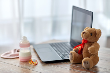 feeding and technology concept - bottle with baby milk formula, laptop computer, teddy bear toy and soother on wooden table at home