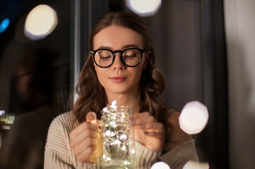 winter, comfort and people concept - young woman in glasses with garland lights in mason jar mug at home