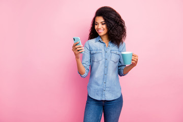 Portrait of positive afro american girl rest relax on spring holidays hold hot caffeine beverage mug use smartphone follow post comment wear denim jeans shirt isolated pink color background