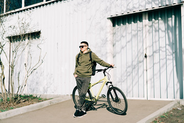 a young man in khaki sportswear holds a Bicycle standing next to him, against the background of a light hangar on a Sunny spring day