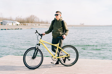 Fototapeta na wymiar a young man in khaki sportswear and glasses stands on a pontoon near the lake and holds a yellow Bicycle near him on a Sunny spring day