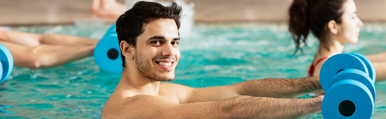 Side view of man smiling at camera while exercising with dumbbells in swimming pool, panoramic shot